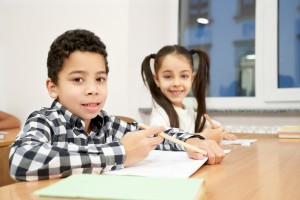A smiling boy and girl sit at a long table pencils and paper.