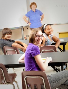 Teacher stands at the front of the room, smiling with her hands on her hips, the middle school students are looking toward the back of the room, smiling at camera. 